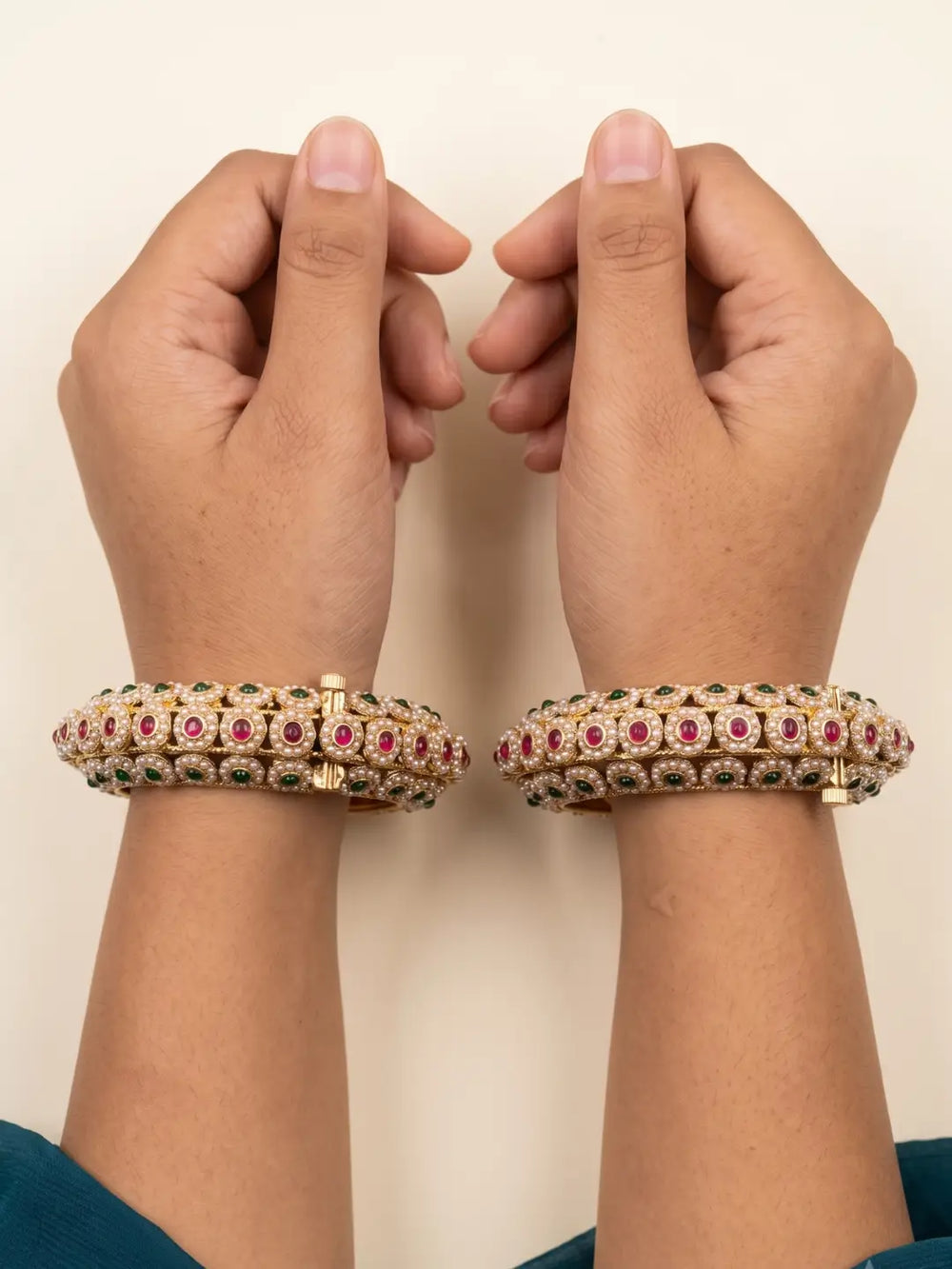 A pair of gold-plated bangle bracelets decorated with red and green stones, worn on two wrists against a plain beige background.