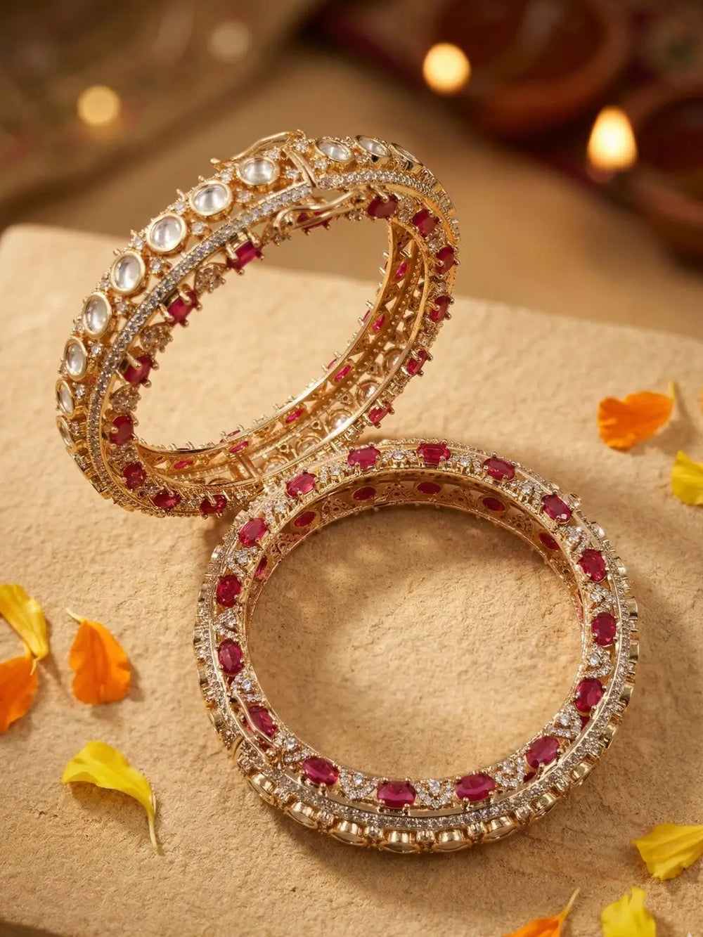 Two ornate gold plated bangles adorned with clear kundan stones and ruby stones, arranged on a beige textured surface with scattered orange and yellow flower petals.