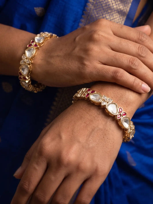 Gold-plated bangles with white polki stones and red floral accents are worn on a person's wrists, shown against a blue fabric background.