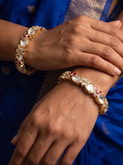 Gold-plated bangles with white polki stones and red floral accents are worn on a person's wrists, shown against a blue fabric background.