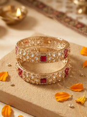 Two stacked gold-plated bangles decorated with white stones and red square accents, displayed on a beige textured block with orange flower petals and a soft warm background.