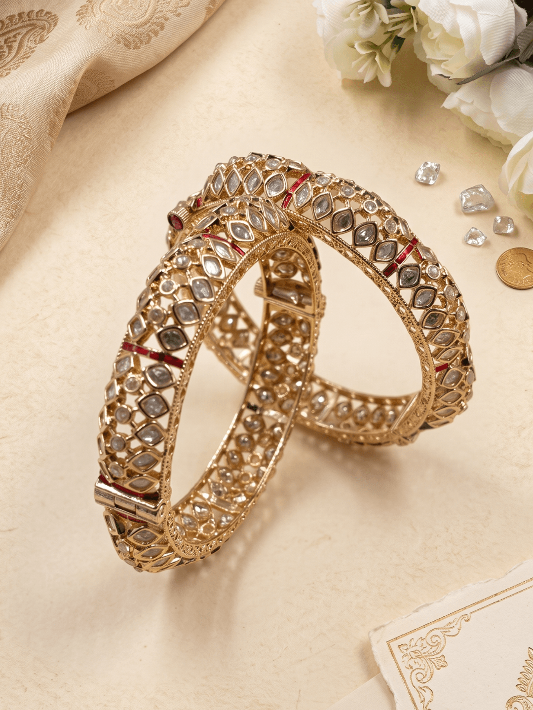 Two ornate gold-toned polki-style bangles decorated with clear stones and small red inlays, arranged on a beige surface with decorative fabric, flowers, and stones around them. Perfect for wedding, festive, and bridal wear.