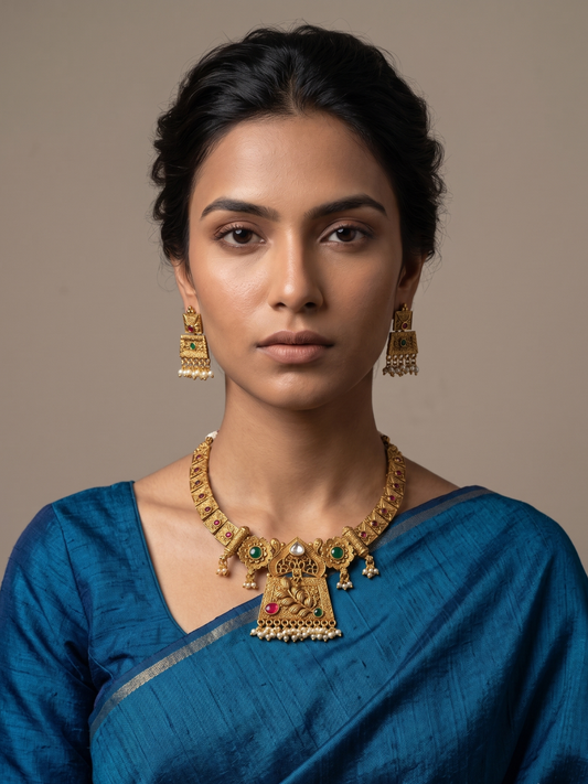 A woman wearing an ornate gold-plated antique gold short necklace and matching earrings with green and red stone accents, paired with a blue saree, against a plain beige background.