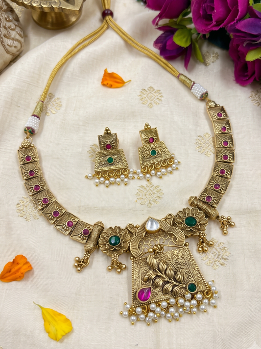 Gold-toned traditional Indian antique jewelry set featuring a necklace and matching earrings with green and pink stones and white pearl accents, arranged on a cream embroidered fabric with decorative flower petals.