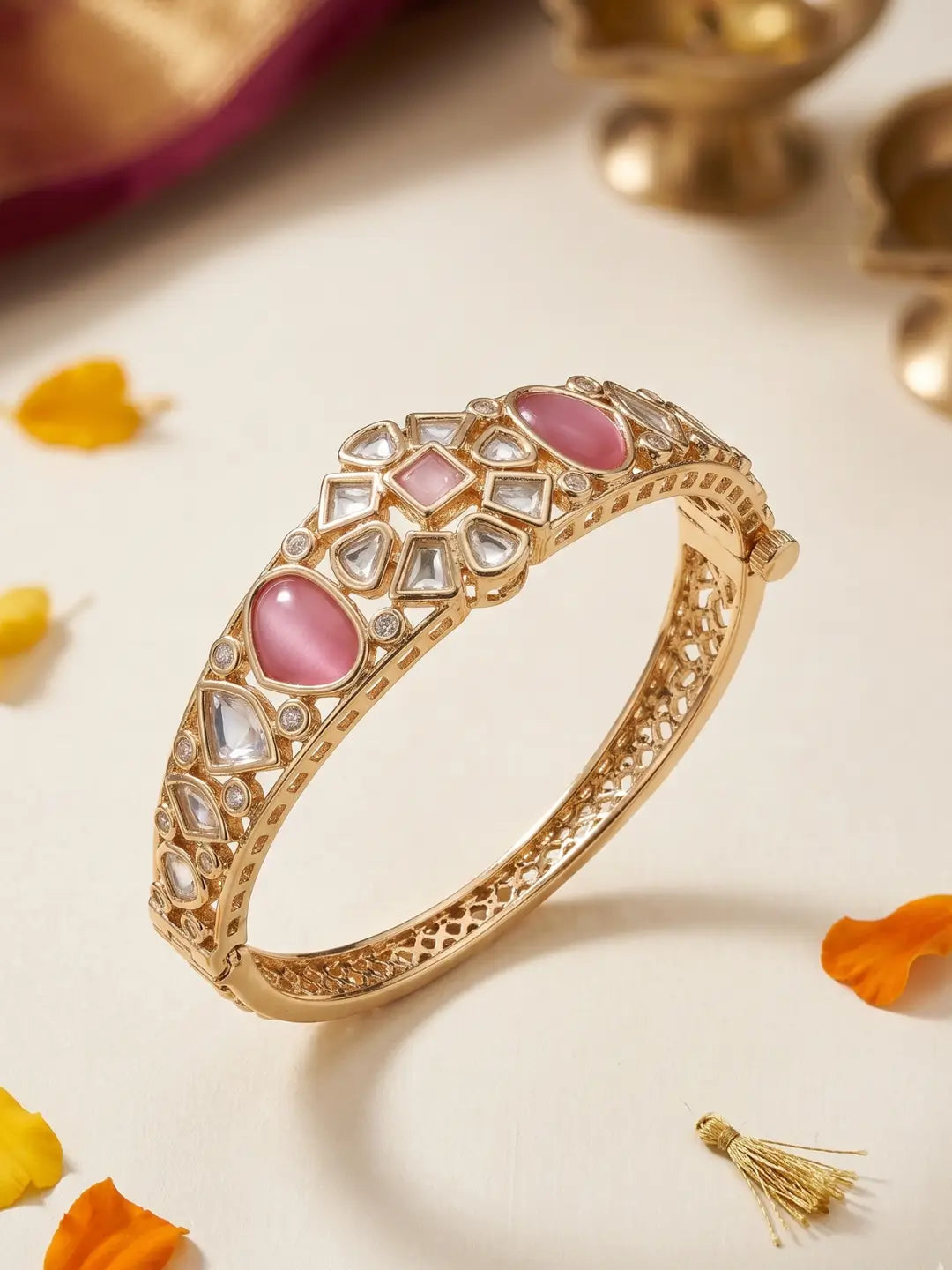 A gold-plated bangle bracelet decorated with pink and clear stones, displayed on a light cream surface with scattered orange flower petals.
