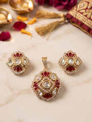 Gold-plated Indian Kundan pendant and earrings set featuring a matching pendant and earrings with red floral inlays and clear stones, displayed on a light marble surface with decorative props.