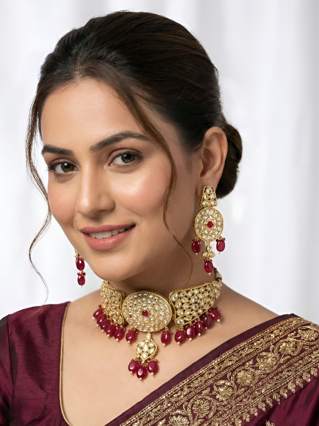 A woman wearing an ornate gold-toned traditional choker necklace with red bead accents and matching drop earrings, paired with a deep maroon sari with gold embroidery.