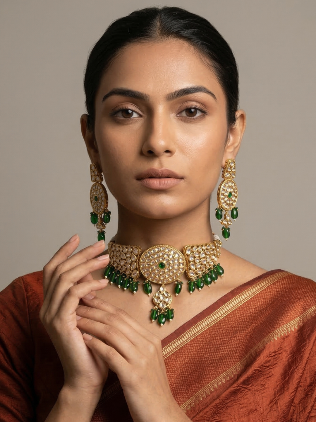 A woman wearing an ornate gold-toned choker jewelry set with green stone accents, including a choker necklace, matching drop earrings, and a rust-colored sari, posed against a neutral beige background.