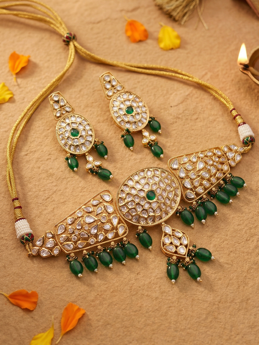 Gold-toned traditional Indian choker jewelry set featuring a necklace, earrings, and a large statement piece, embellished with white stones and green beads, arranged on a textured beige surface with scattered orange and yellow petals.