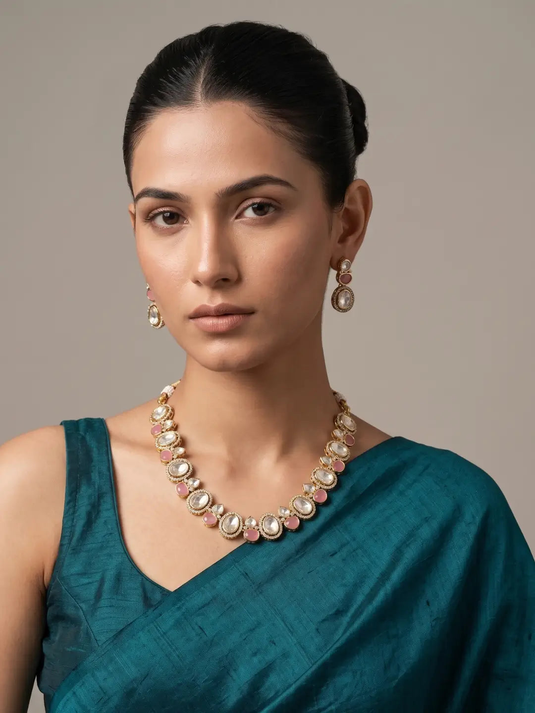 A woman wearing a gold-toned statement kundan necklace with pink and white stones, matching drop earrings, and a teal green sari against a neutral beige background.