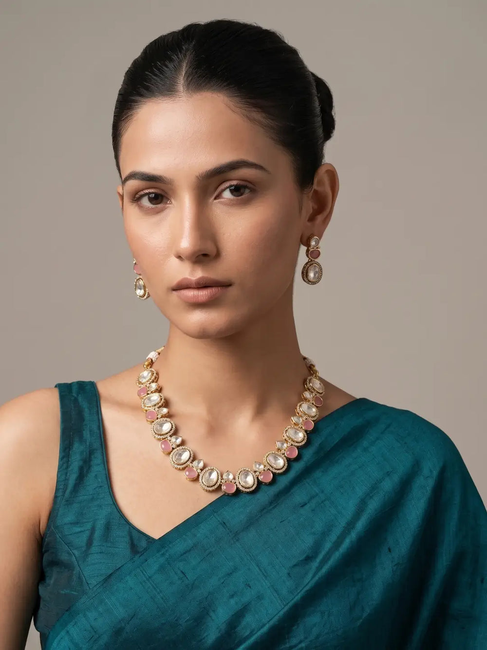 A woman wearing a gold-toned statement kundan necklace with pink and white stones, matching drop earrings, and a teal green sari against a neutral beige background.