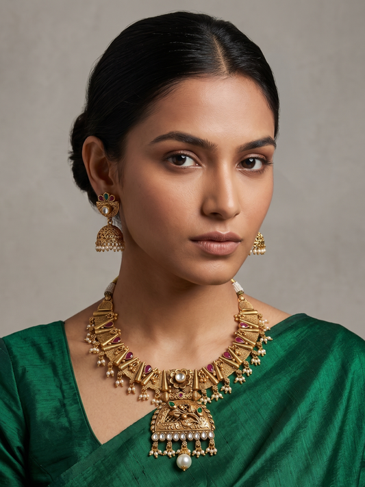 A woman wearing an ornate, antique gold-toned necklace with matching earrings and a green blouse, photographed against a soft, neutral background.