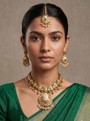 A woman wearing traditional Indian antique kundan jewelry, including a gold-toned maang tikka, matching earrings, and a statement necklace, paired with a green saree against a neutral beige background.