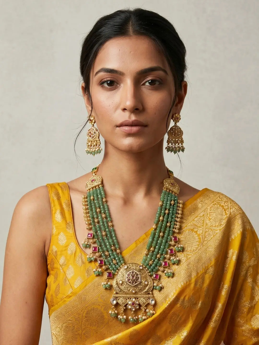 A woman wearing a traditional Indian yellow saree and a large, ornate green-and-gold beaded necklace with matching earrings, set against a neutral beige background.