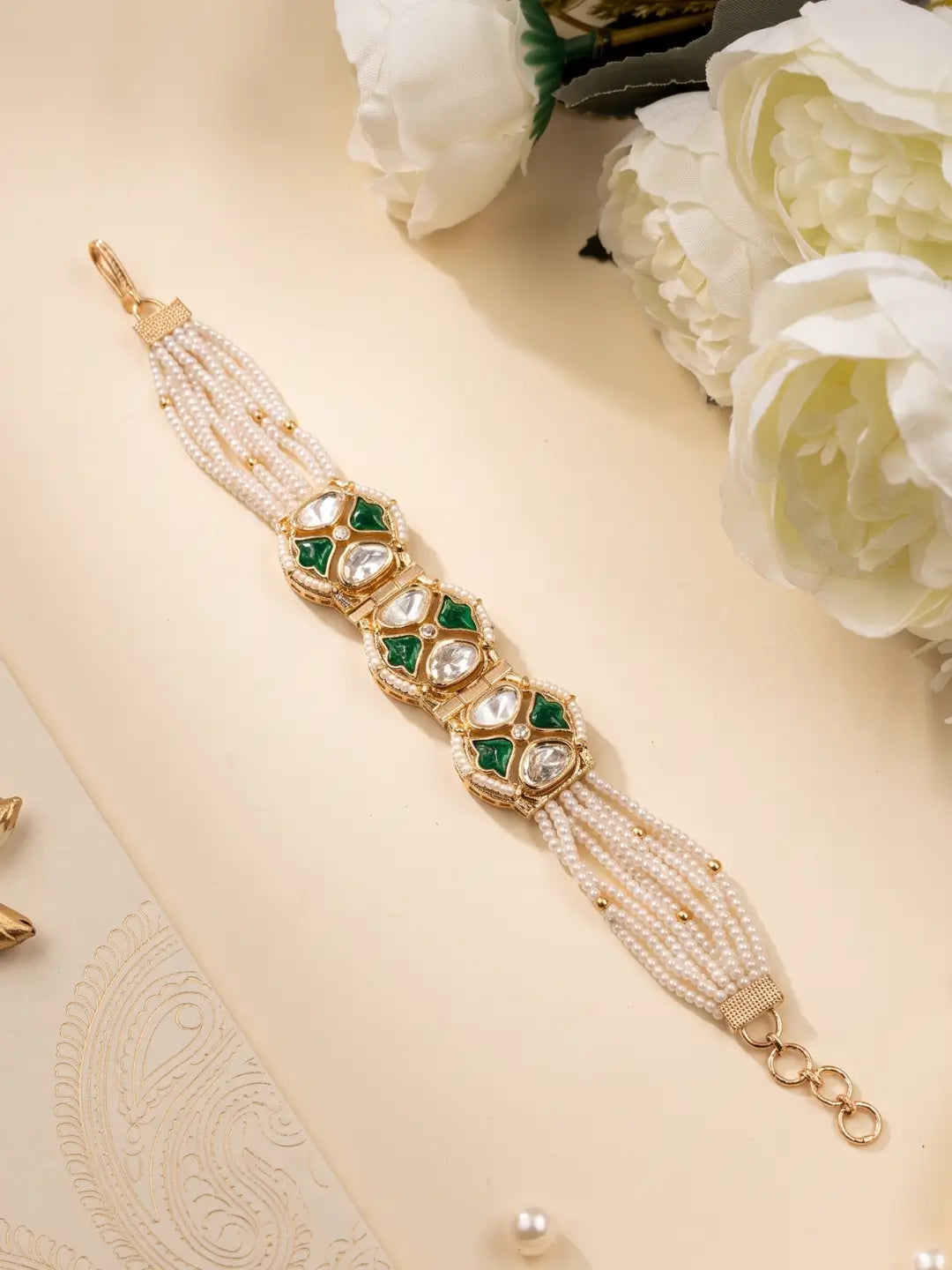 A gold-plated multi-strand pearl bracelet with green meena and clear polki stone embellishments, styled on a beige surface with white flowers.