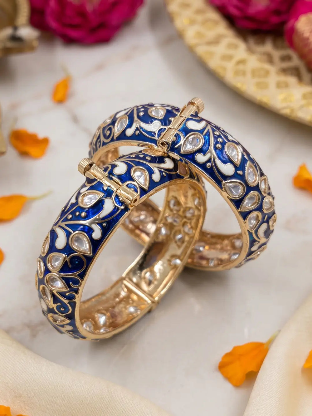 Two gold-plated bangles decorated with intricate blue and white enamel patterns and small clear stones, displayed on a light marble surface with decorative orange petals and flowers.