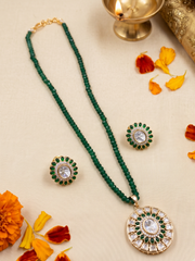A green beaded necklace with a round kundan pendant and matching stud earrings, styled on a cream fabric background with orange flower petals and a brass diya.
