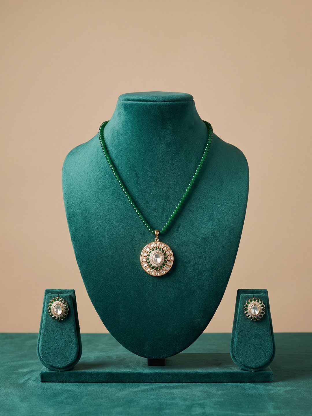 A jewelry set displayed on a teal velvet mannequin bust, featuring a beaded necklace with a round kundan pendant and matching stud earrings on a beige background.