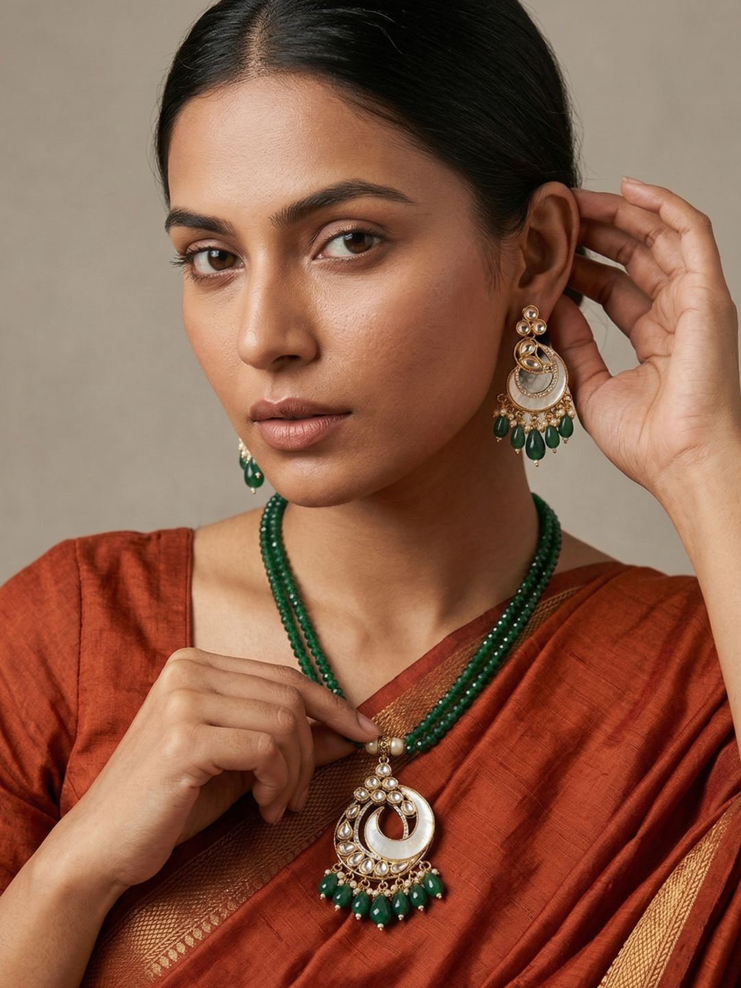 A woman wearing a green beaded mother of pearl and kundan necklace and matching drop earrings with gold-toned circular details, styled with a rust-colored sari against a neutral beige background.