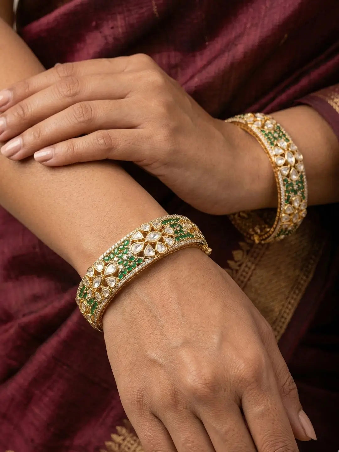 A close-up of a person wearing two ornate gold bangles decorated with green stones and clear stones, set against a deep maroon fabric background.