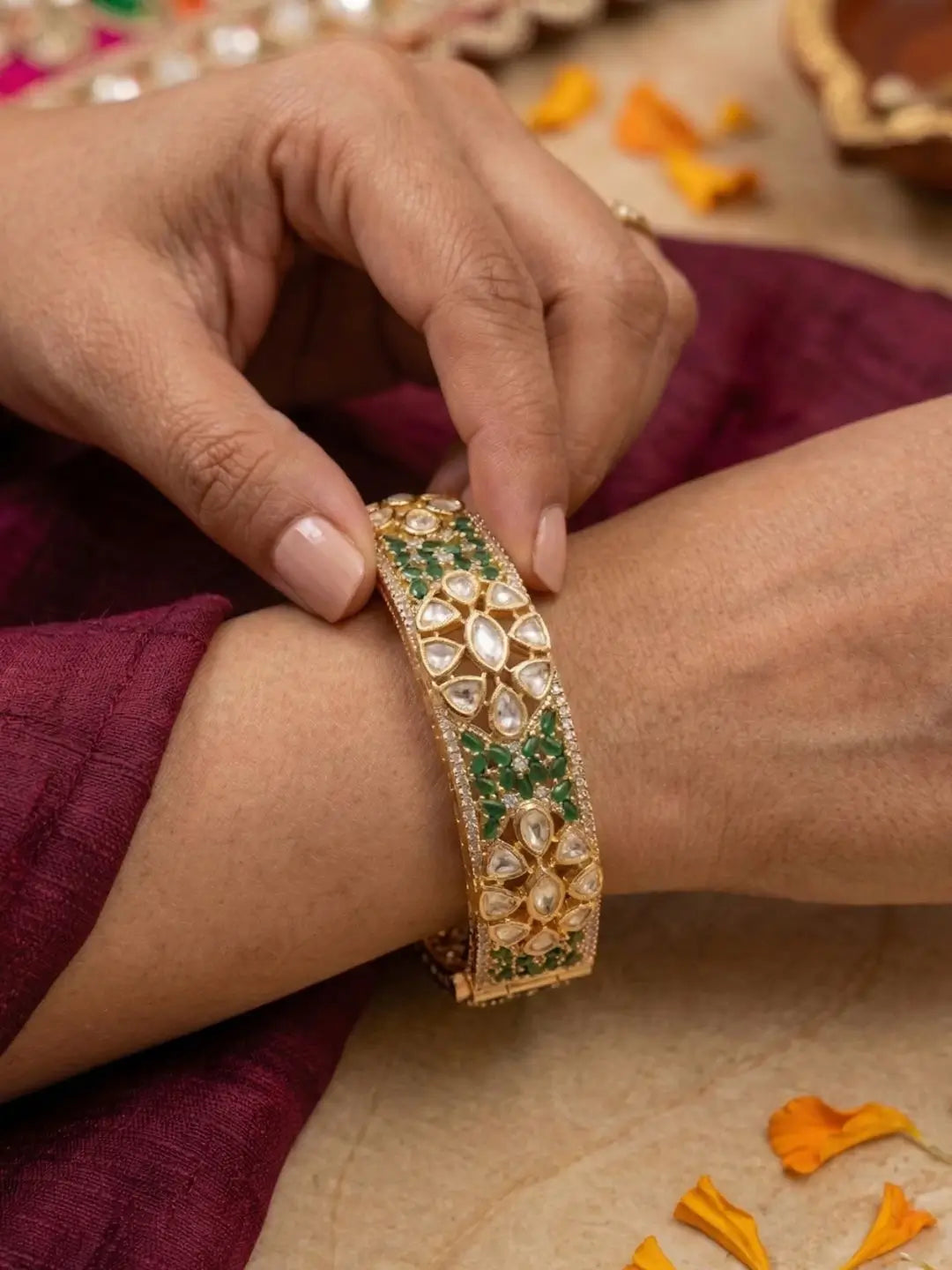 Person wearing a gold bracelet with green stones on a maroon fabric background.