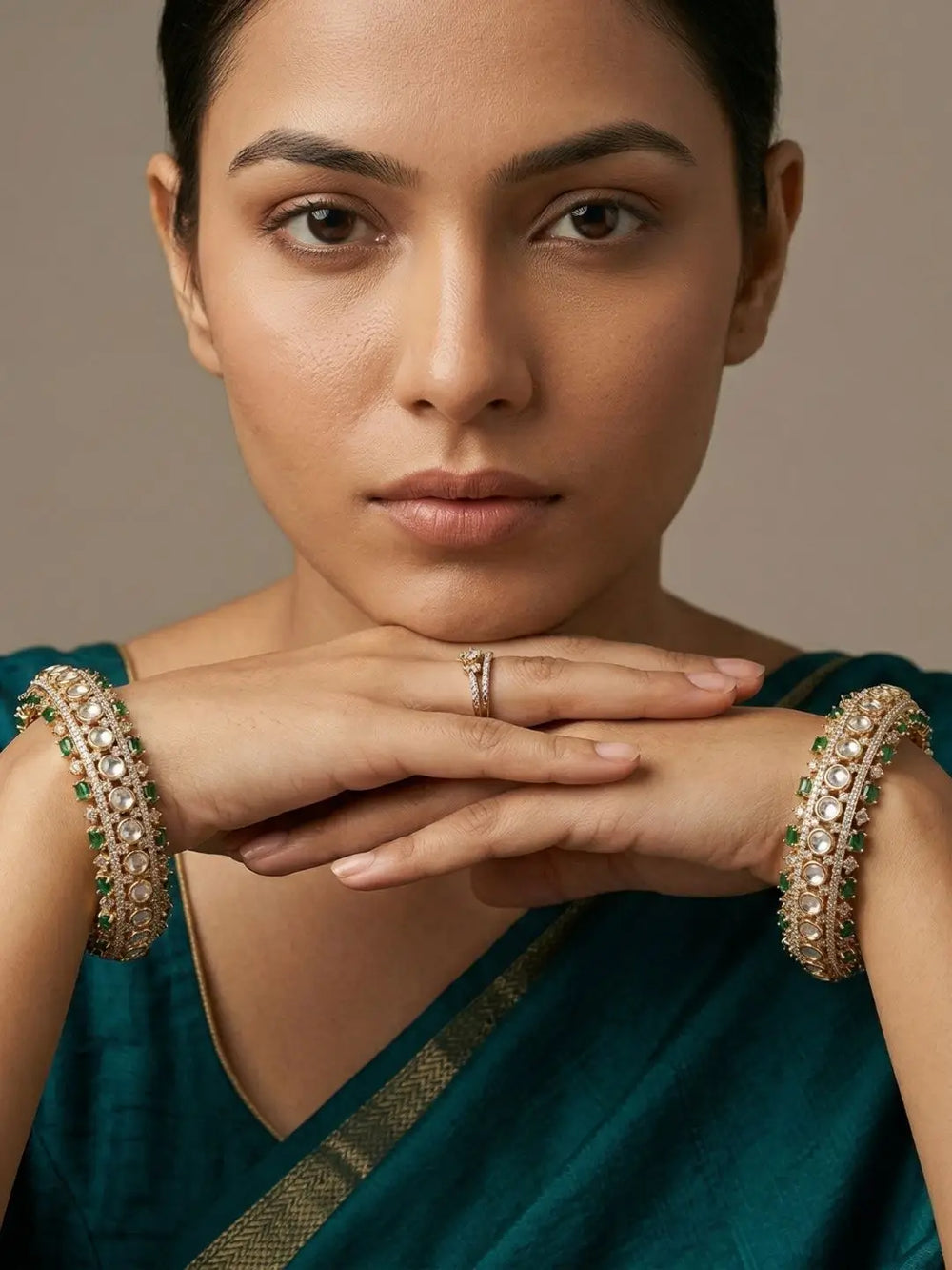 A close-up portrait of a woman wearing ornate gold-toned bangles with green and white stone embellishments on both wrists, along with a matching ring on her finger. She is wearing a deep teal sari against a soft neutral background.