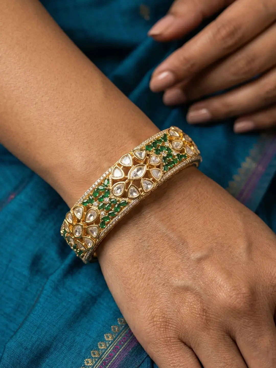 A close-up of a person's wrist wearing an ornate gold-toned bangle bracelet decorated with green stones and clear kundan stones, set against a teal blue fabric background.