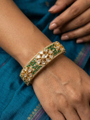 A close-up of a person's wrist wearing an ornate gold-toned bangle bracelet decorated with green stones and clear kundan stones, set against a teal blue fabric background.