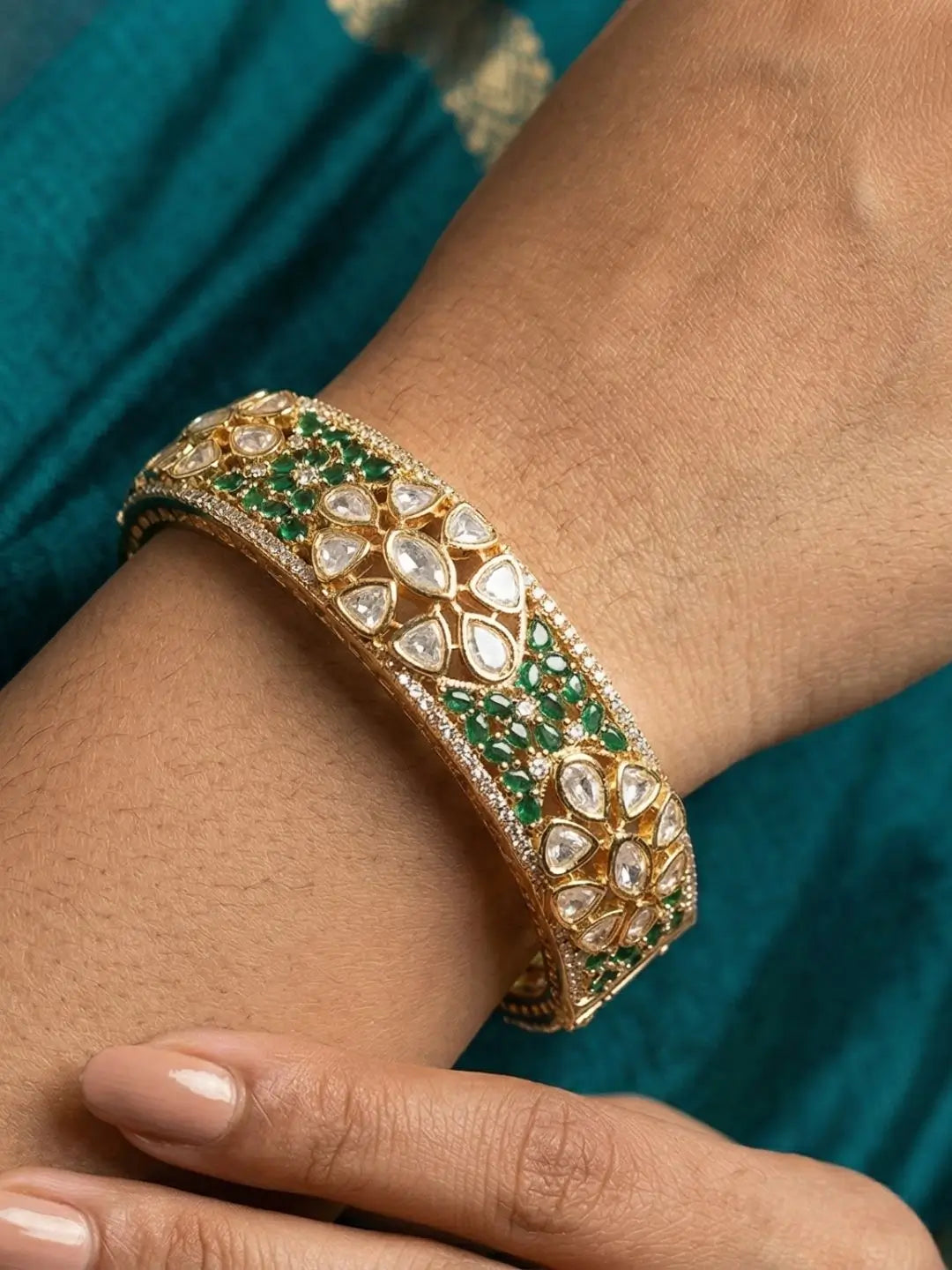 Gold bracelet with green and white polki stones on a wrist against a teal fabric background