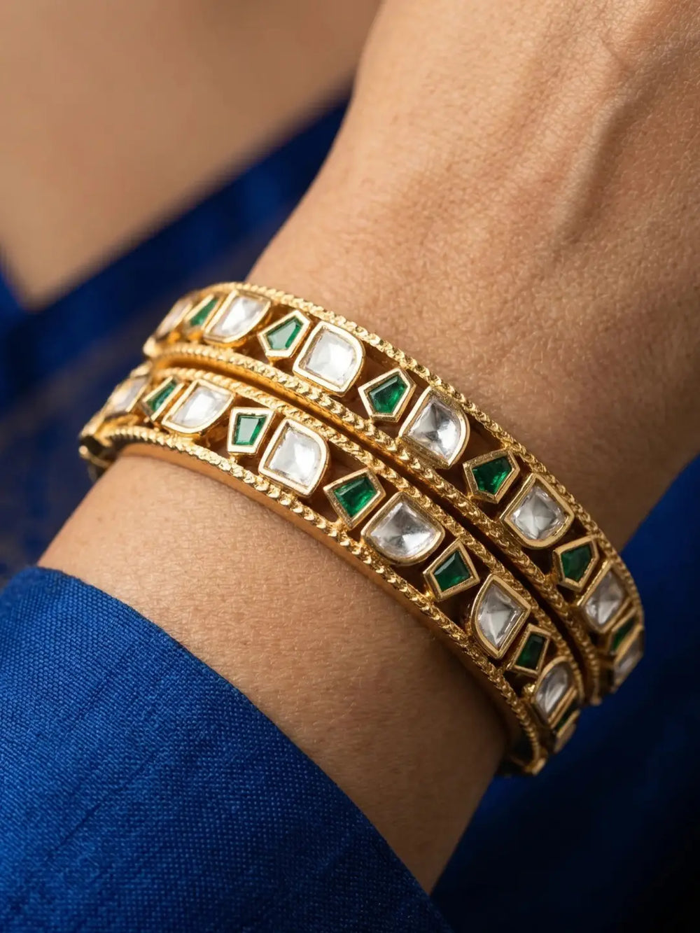 A close-up of a person's wrist wearing three stacked gold bangles decorated with white stones and green inlay.