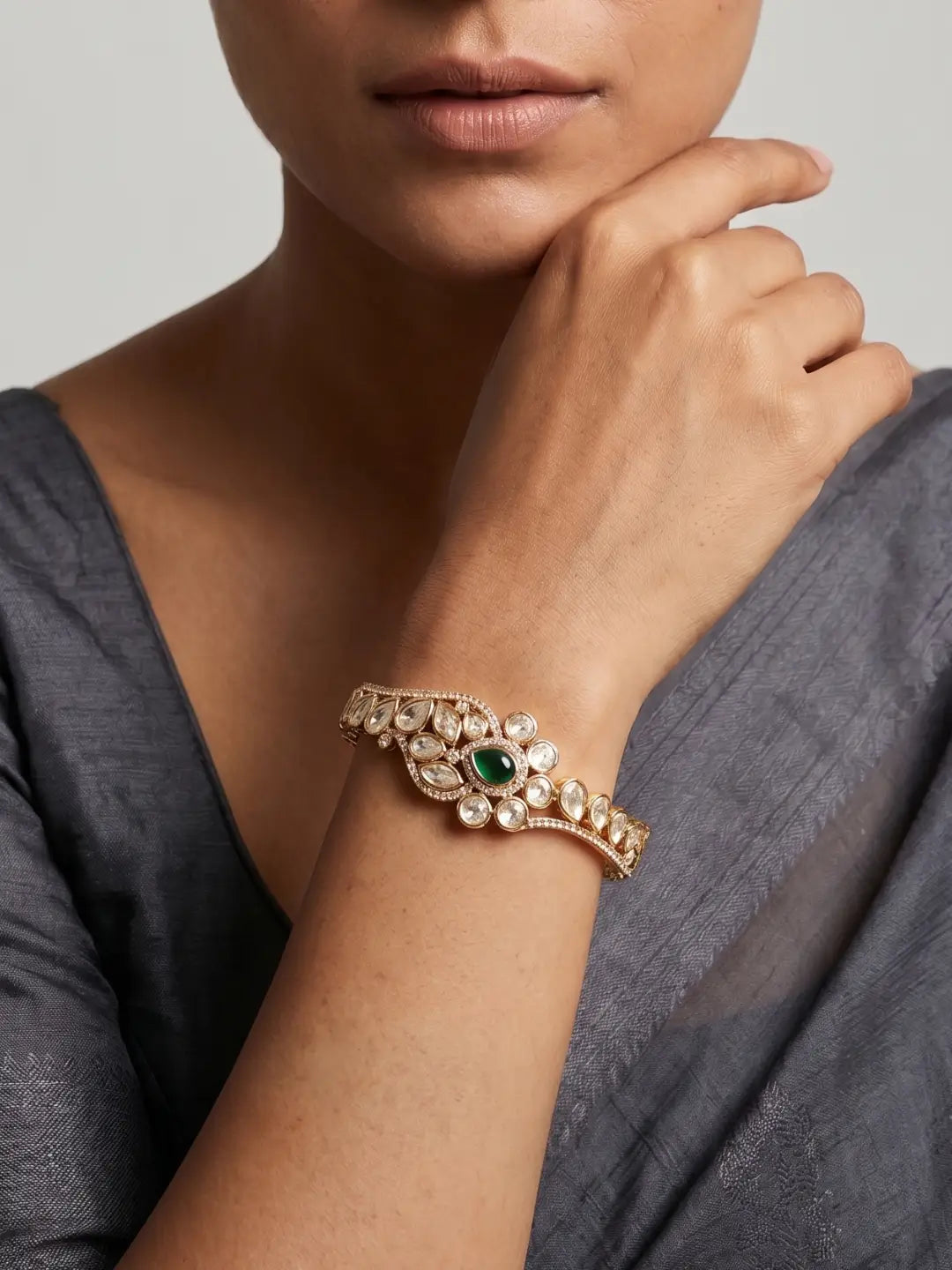 A close-up of a woman wearing a gold-toned ornate bracelet with a green oval stone and clear stones, shown on her wrist against a soft gray background.