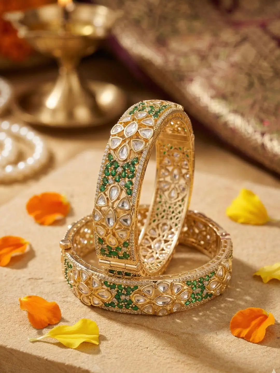 Ethnic gold-plated bangles studded with green stones, polki and CZ Stones, placed on a textured surface with orange and yellow flowers.