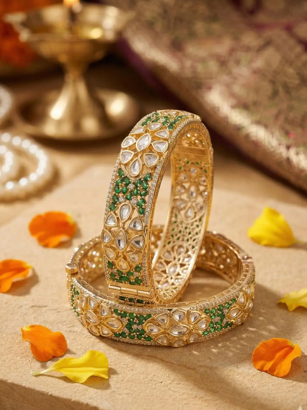 Ethnic gold-plated bangles studded with green stones, polki and CZ Stones, placed on a textured surface with orange and yellow flowers.