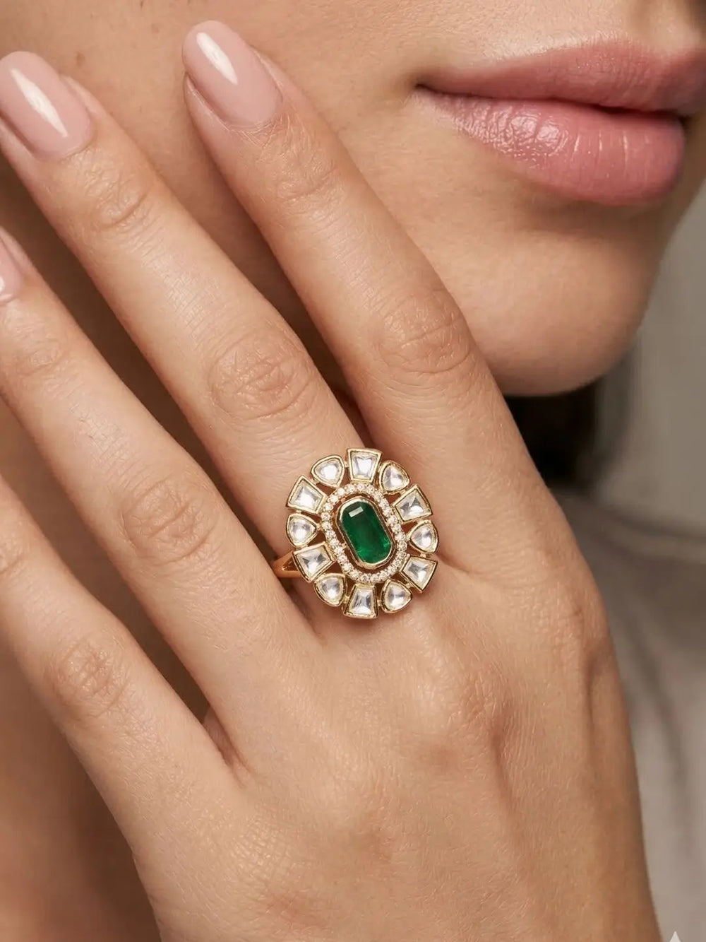 A close-up of a woman's hand wearing a gold ring with a central green emerald-cut stone surrounded by small clear stones.