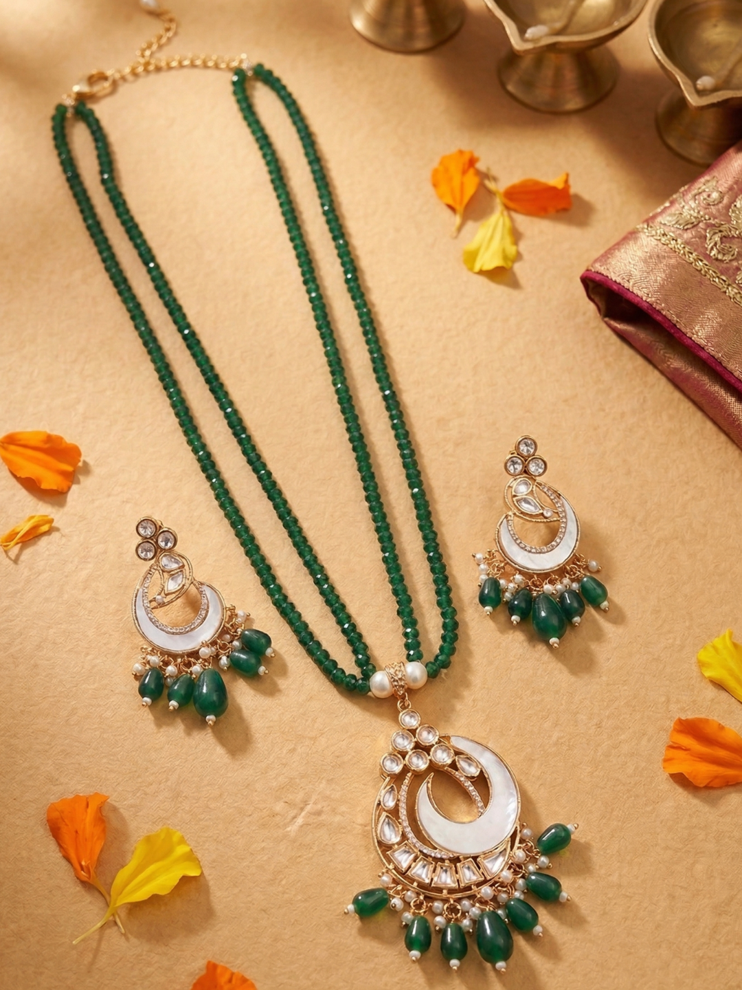 A traditional Indian jewelry set featuring a double-strand green beaded necklace with a large crescent moon mop pendant, matching green and white chandelier earrings, and a gold chain, arranged on a beige surface with decorative orange and yellow petals.