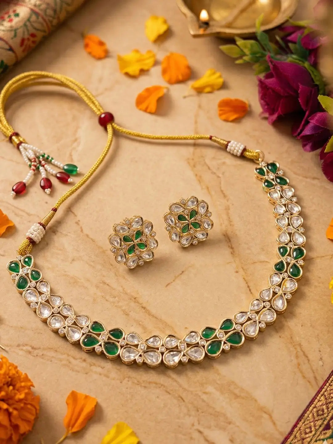 Gold-tone Indian kundan jewelry set featuring a necklace and matching earrings with green stones and clear polki accents, arranged on a beige marble surface with decorative flowers and petals.