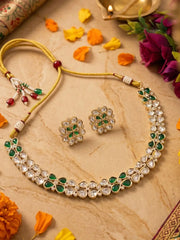 Gold-tone Indian kundan jewelry set featuring a necklace and matching earrings with green stones and clear polki accents, arranged on a beige marble surface with decorative flowers and petals.