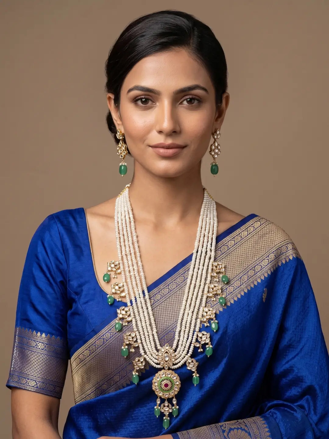 A woman in a blue saree is wearing an Elegant multi-layer pearl long necklace set featuring a handcrafted Kundan pendant with emerald green bead drops, ruby-inspired centre stone, and matching earrings, perfect for bridal wear, weddings, festive occasions, and traditional Indian jewellery.