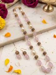 A pink beaded jewelry set featuring a long necklace and matching earrings with light pink stone beads, white faux pearls, and ornate gold-toned filigree spacer beads, styled on a marble slab with flowers and petals.