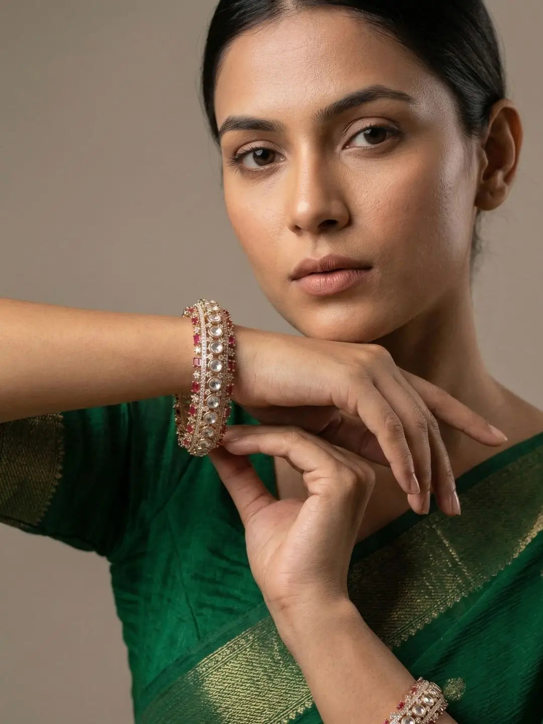 A close-up portrait of a woman wearing a green sari and matching green jewelry, with one hand resting on her chin while wearing a gold-toned bangle decorated with red and white stones.