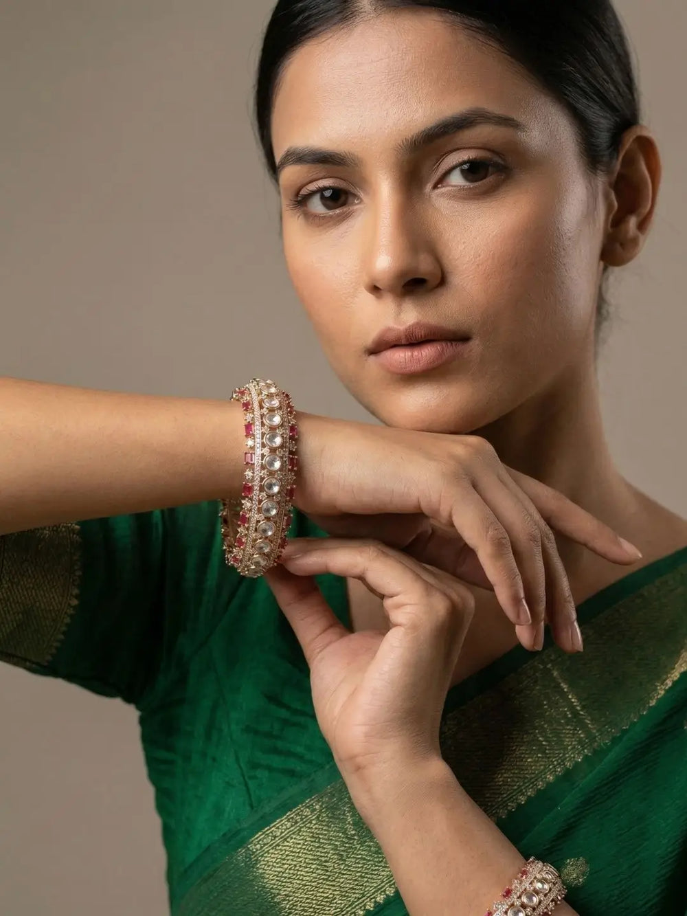 A close-up portrait of a woman wearing a green sari and matching green jewelry, with one hand resting on her chin while wearing a gold-toned bangle decorated with red and white stones.