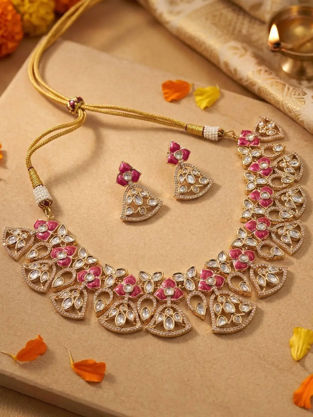 Gold-toned Indian bridal jewelry set featuring a heavily embellished necklace and matching earrings with pink and white stones, displayed on a beige textured surface with decorative flower petals.