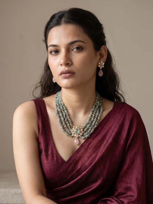 A woman wearing a deep burgundy draped blouse and a layered sage green beaded necklace with a gold-toned ornate pendant, paired with matching earrings, set against a plain beige background.