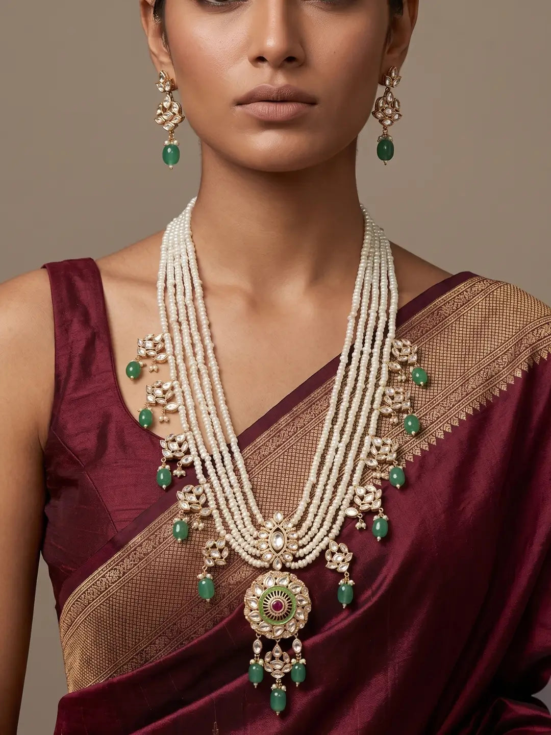 A woman in  red saree is wearing an Elegant multi-layer pearl long necklace set featuring a handcrafted Kundan pendant with emerald green bead drops, ruby-inspired centre stone, and matching earrings, perfect for bridal wear, weddings, festive occasions, and traditional Indian jewellery.