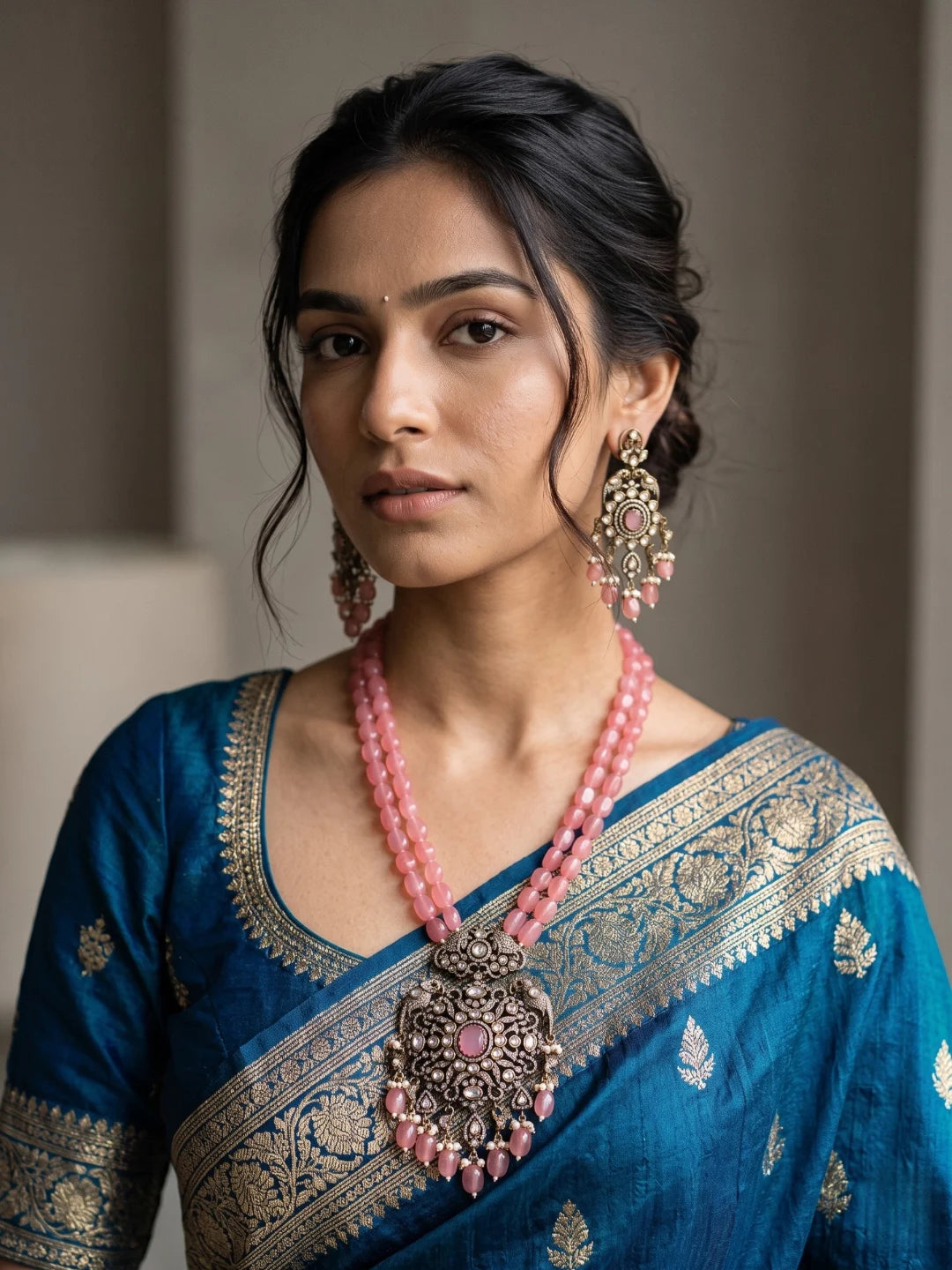 A woman wearing a blue saree with gold zari embroidery, a pink beaded antique Victorian moissanite polki necklace with a large ornate pendant with a parrot motif, and matching pink and gold long drop earrings, shown in a close-up portrait.