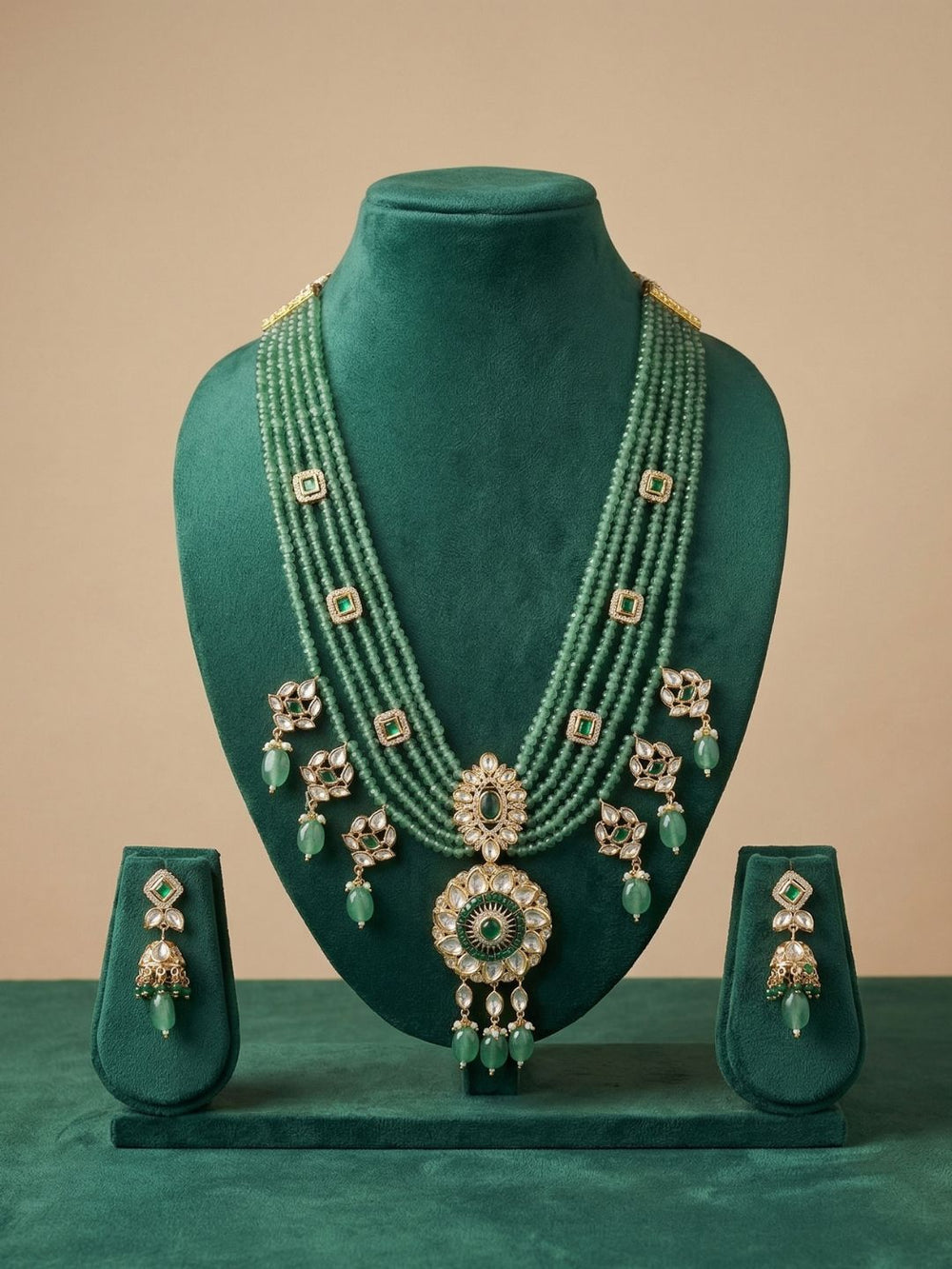 A traditional Indian jewelry set displayed on a dark green velvet mannequin bust, featuring a multi-strand green beaded necklace with a large central pendant, matching green beaded earrings, and a small green beaded drops, all set against a solid beige background.
