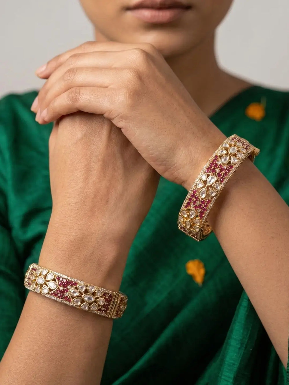 A close-up of a woman wearing two ornate gold-plated bangles with studded ruby and polki kundan stones, paired with a green embroidered garment.