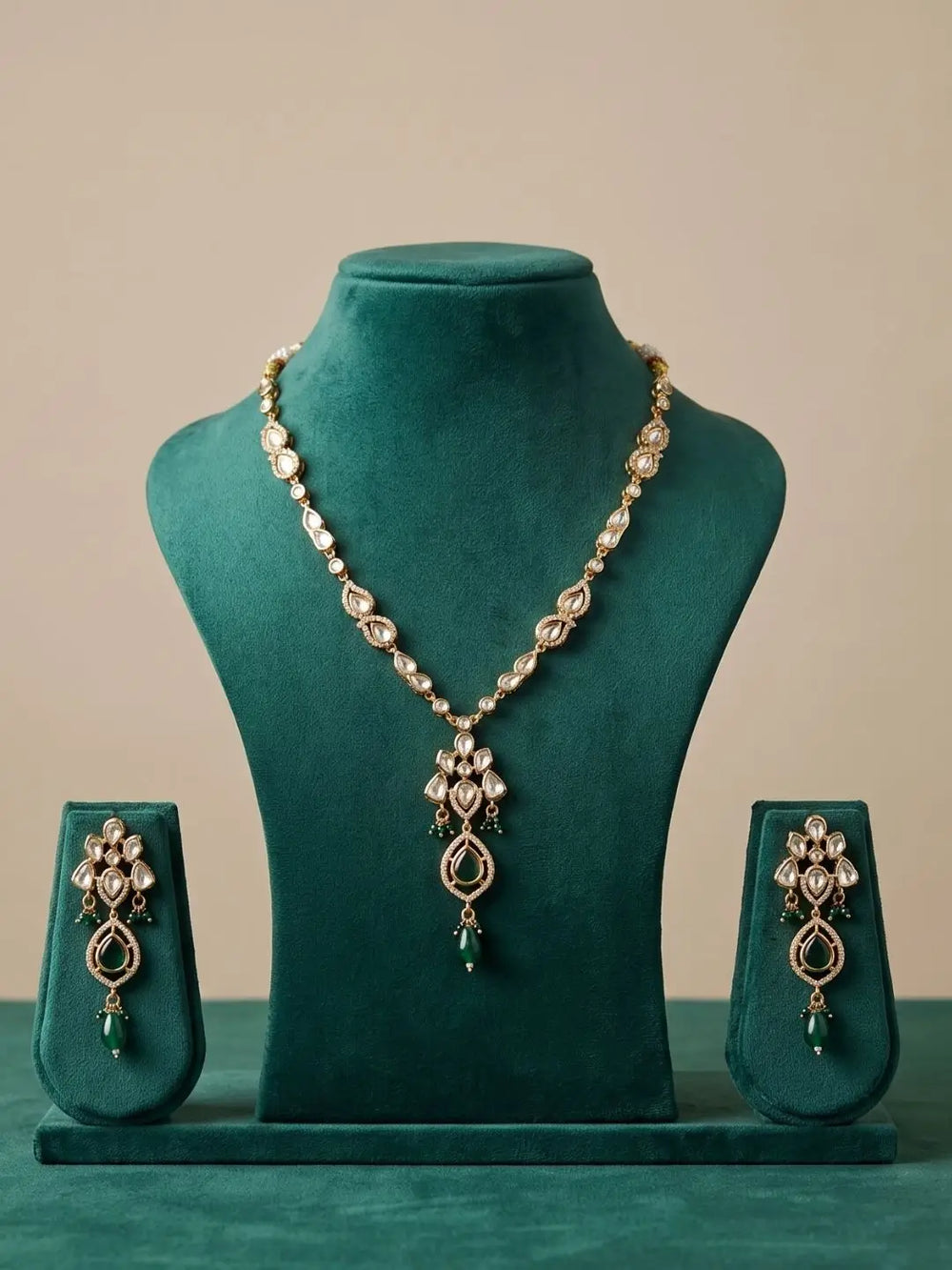 Gold-plated polki pendant jewelry set featuring a necklace and matching earrings with clear stones and green teardrop accents, displayed on a dark green velvet jewelry stand.