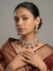 A woman wearing a traditional Indian choker necklace and matching earrings with red and gold elements, paired with a maroon and gold saree, set against a plain gray background.