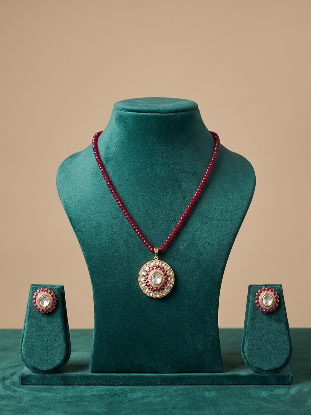 A red beaded necklace with a round kundan pendant and matching stud earrings displayed on a teal velvet jewelry stand against a beige background.